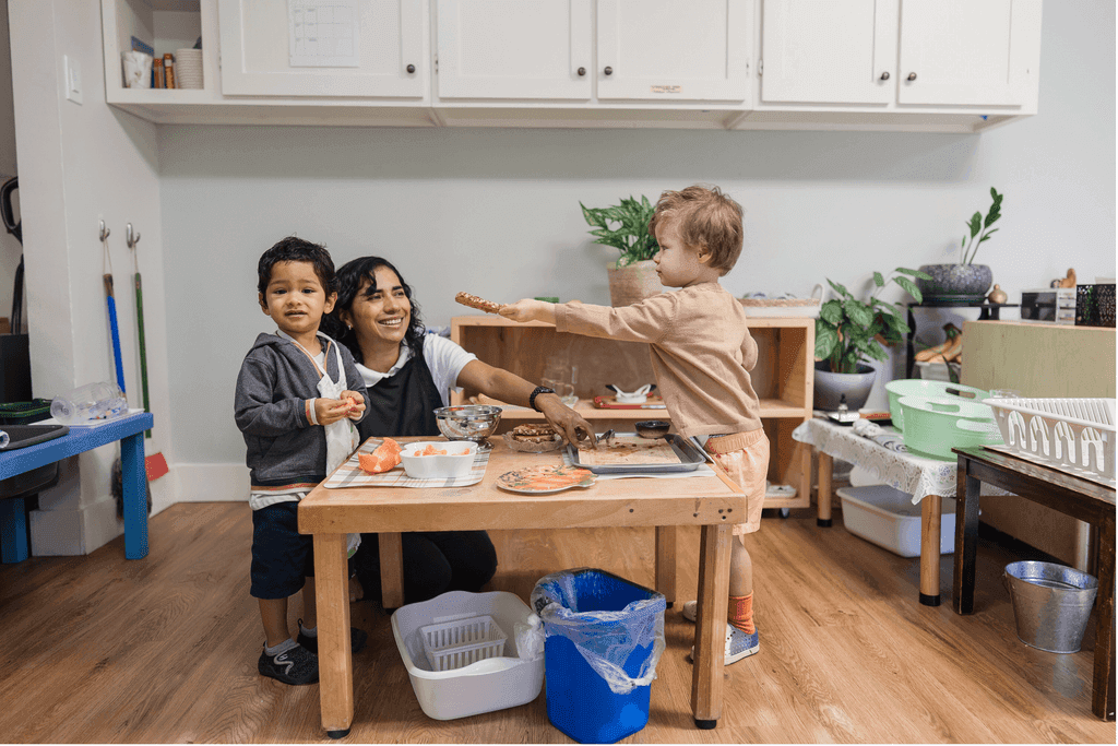 A guide helps two toddlers with a food-preparation activity at a low table